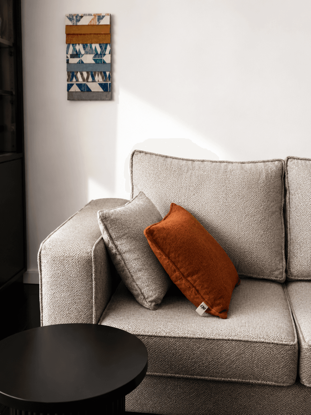 A rust velvet cushion on a greige weave NDB sofa, beneath geometric wall art.