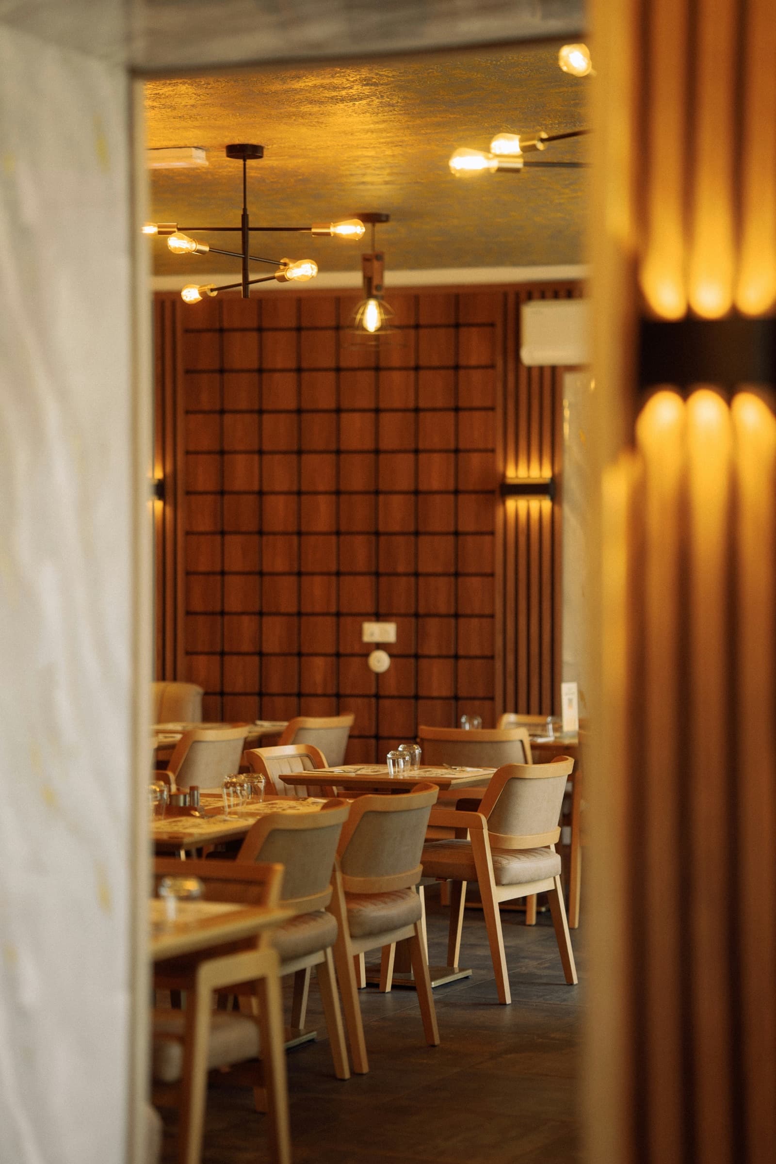 A warm restaurant dining room with timber grid panelling, brass pendants and wooden armchairs.